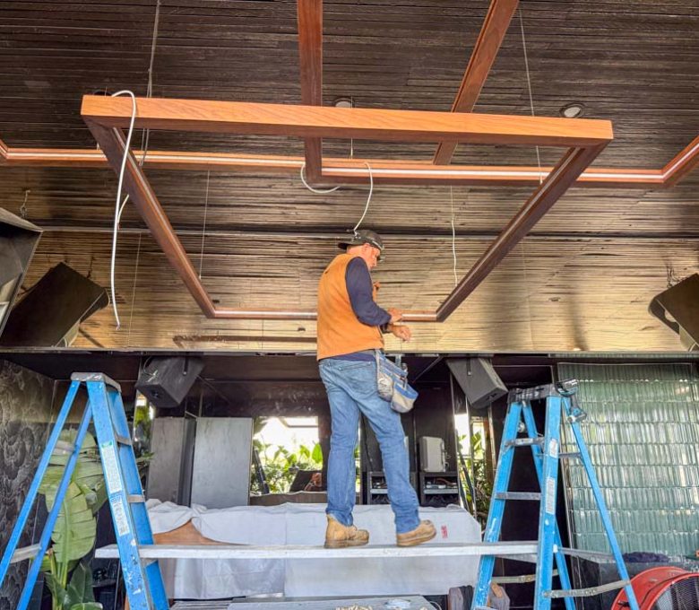 An electrician from Mizo Electric on scaffolding, carefully hanging large, custom-made wooden light fixtures in a restaurant with a dark wood-plank ceiling, creating a stunning focal point for the dining area.