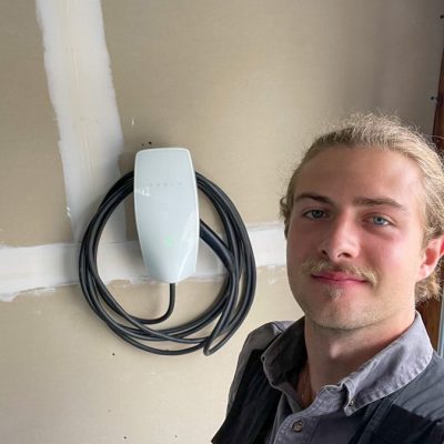 A professional electrician standing next to a newly installed Tesla EV charger, promoting eco-friendly energy solutions.