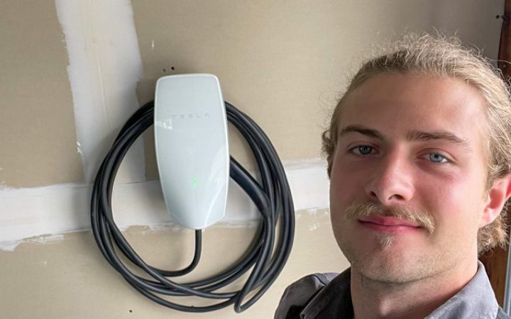 A professional electrician standing next to a newly installed Tesla EV charger, promoting eco-friendly energy solutions.