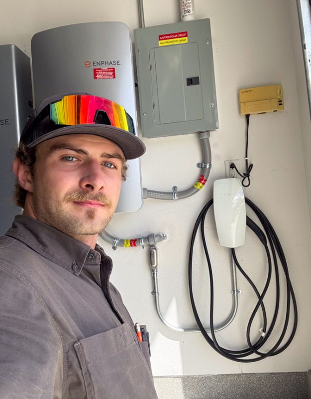 A Mizo Electric technician posing in front of a clean installation featuring an Enphase solar microinverter system and a Tesla Wall Connector, providing homeowners with a complete renewable energy and charging solution.